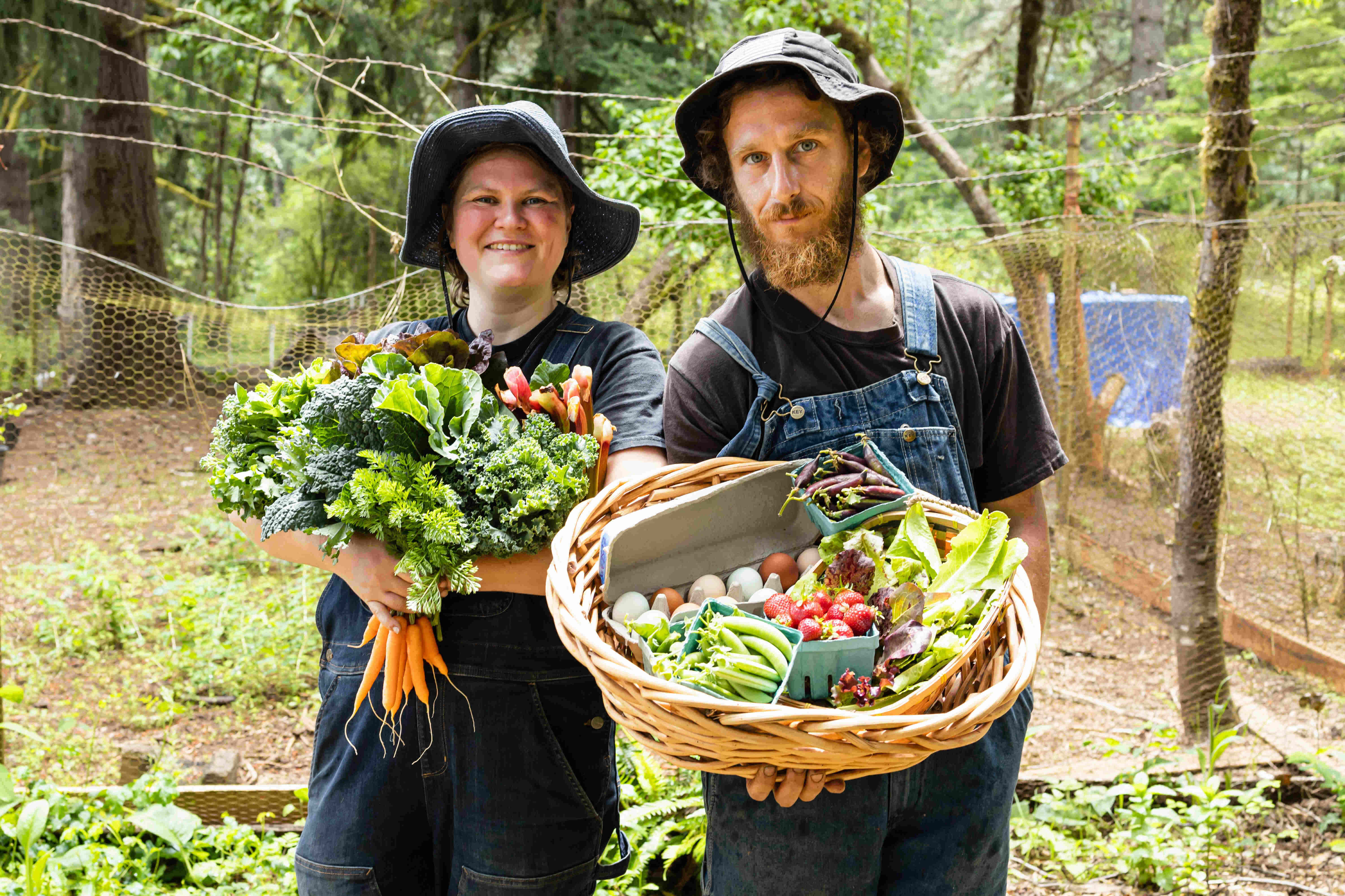 Farmers Cody and Linea Stand next to each other. Cody holds a basket filled with vegetables, eggs, and strawberries. Linea has her arm full of bundles of Kale and a dangling bunch of carrots.